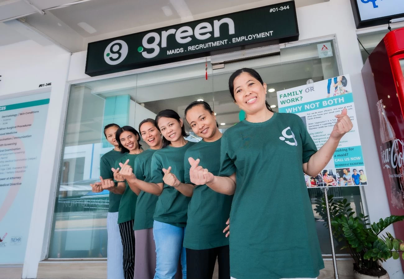 Group of smiling domestic helpers in green shirts posing outside Green Employment maid recruitment agency.