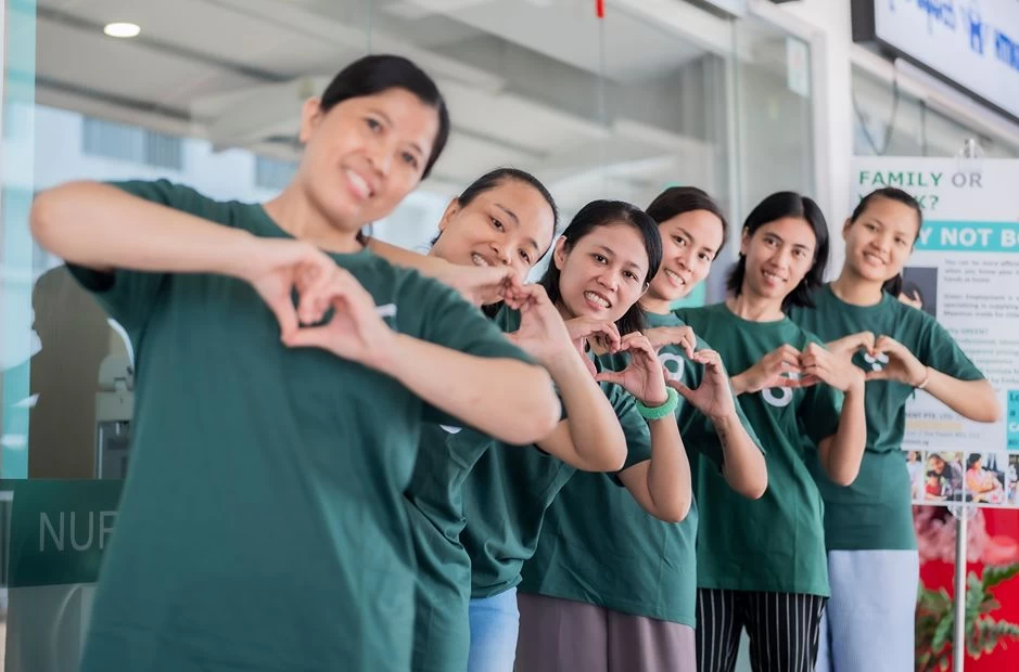 A group of domestic helpers in green shirts makes heart gestures outside the Green Employment office.